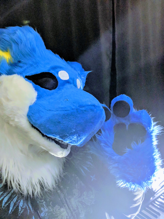 Close-up of a bright blue and white manokit fursuit head raising a blue paw toward the camera