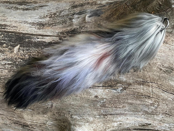 Long gray, black, and white yarn wolf tail with a metal attachment ring, displayed on weathered wood