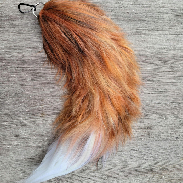 Fluffy orange and brown yarn fox tail with a white tip and metal clip, displayed on gray wood