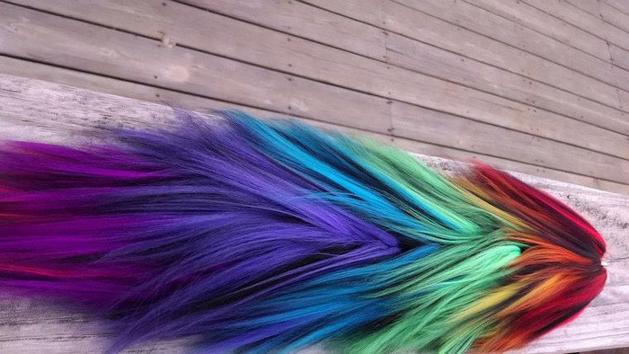 Long fluffy yarn tail blending through red, orange, green, blue, purple, and magenta on weathered wood