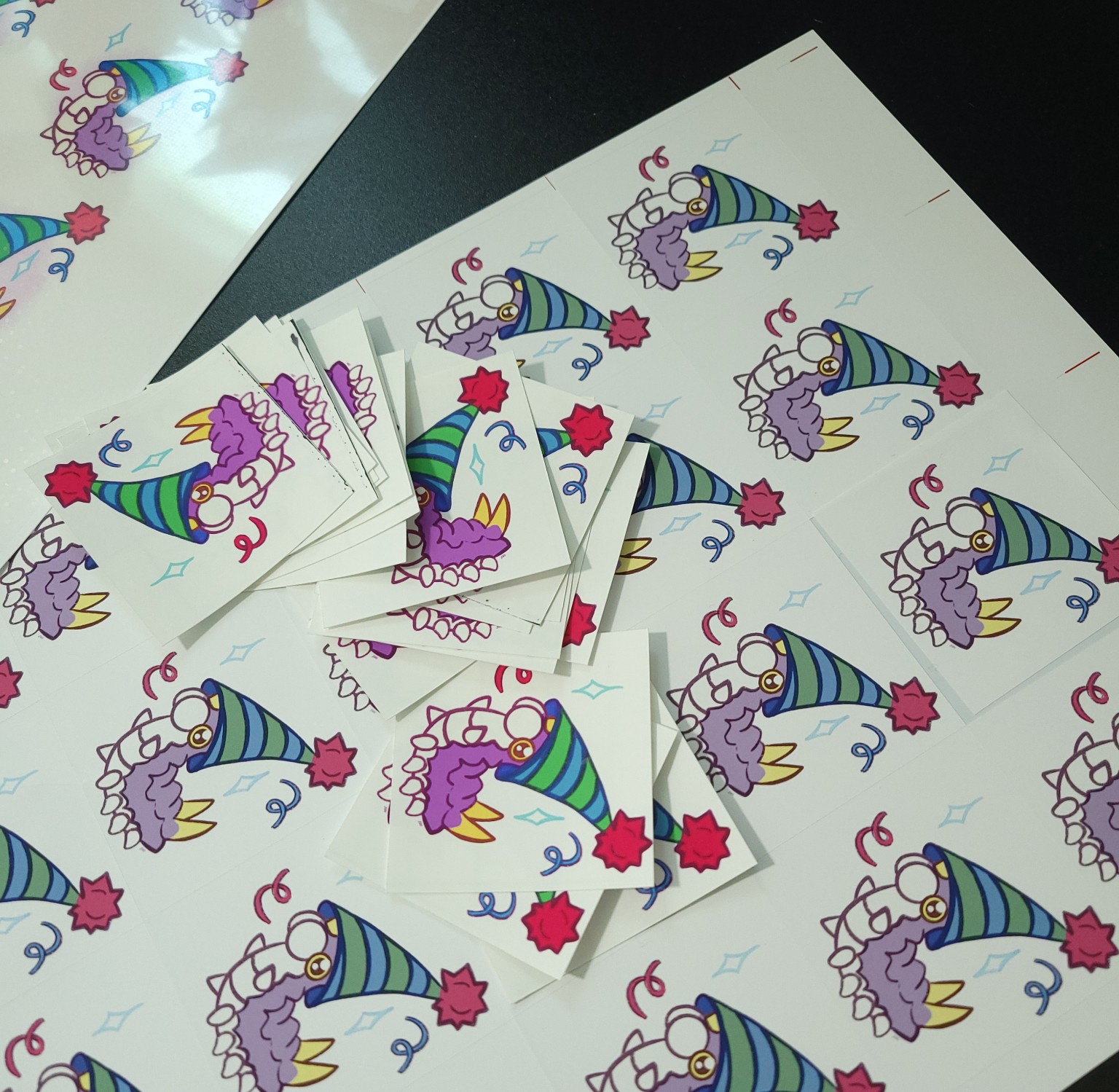 A tabletop covered in party Wurmple stickers, with stacks of individually cut stickers scattered across several printed sheets. This colorful Wurmple graveyard is a batch of misprints that didn't make the cut after color and print quality issues were discovered.