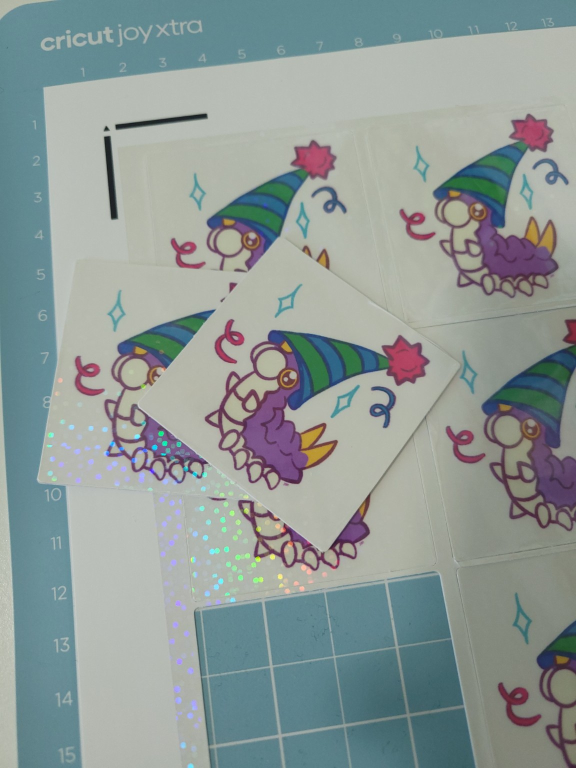 A completed batch of party Wurmple stickers on a Cricut Joy Xtra cutting mat, with one finished sticker displayed in the center. The neatly cut edges and colorful artwork celebrate the end of a trial-and-error journey from misprints to a successful sticker.